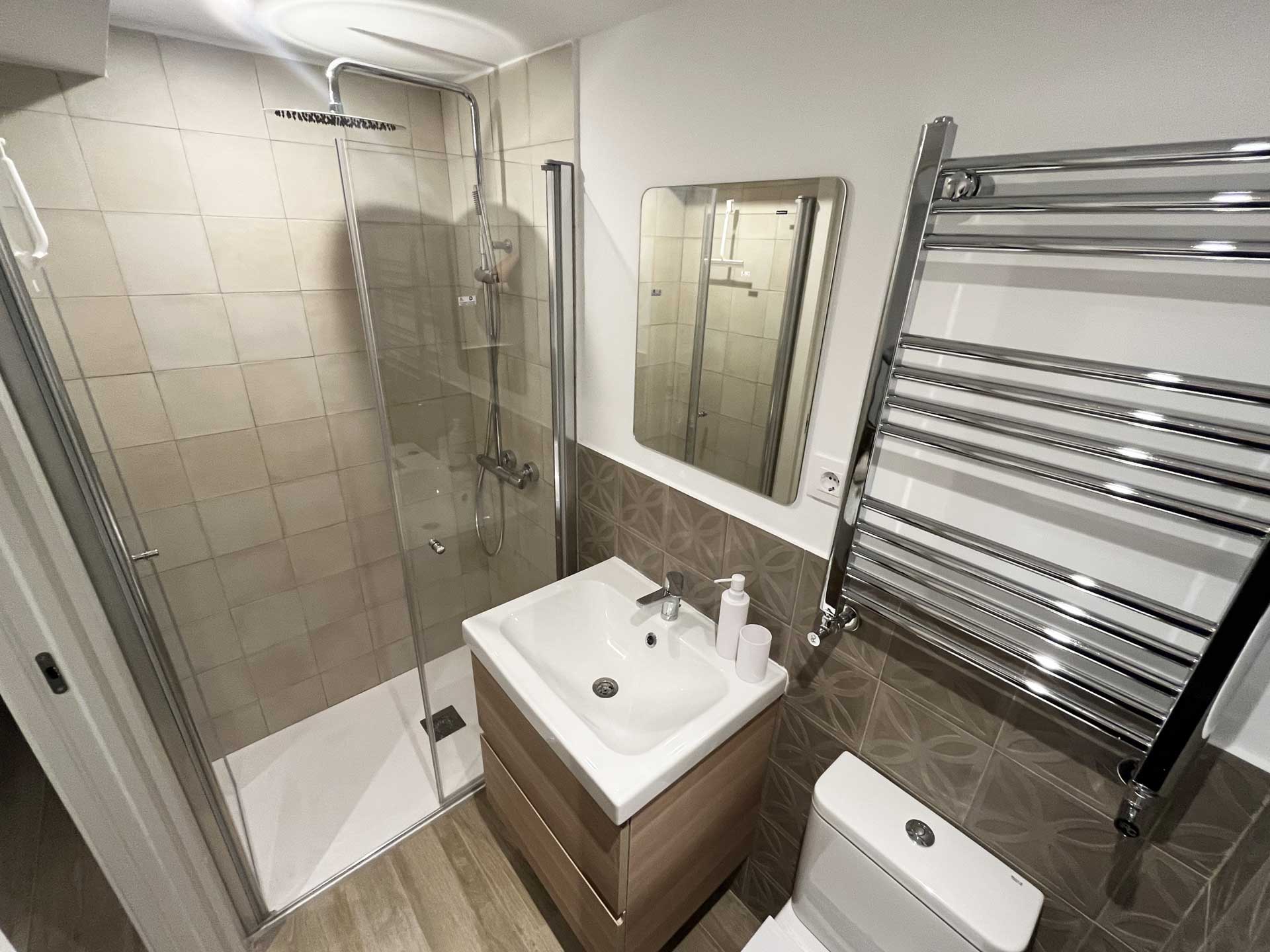 Baño de la habitación de invitados en planta sótano con plato de ducha con vidrieras sin perfiles, lavabo blanco sobre mueble de madera suspendido e inodoro. Realizado por la empresa de reformas integrales en Pozuelo honrados