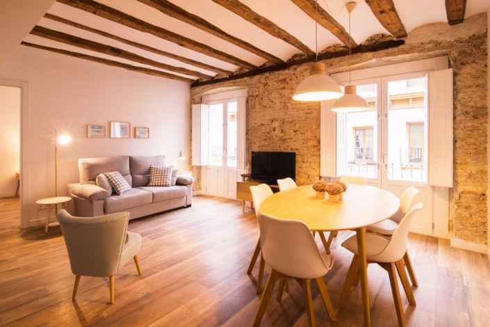 Salón comedor reformado en Madrid que fusiona historia y modernidad. Destacan las vigas de madera originales del techo y una pared de ladrillo visto restaurado. El espacio cuenta con un sofá contemporáneo, una mesa de comedor de madera clara con sillas modernas y una iluminación cálida, reflejando la esencia de la vivienda antigua con toques de interiorismo actual.