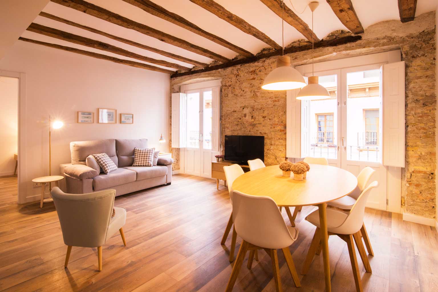 Salón comedor reformado en Madrid que fusiona historia y modernidad. Destacan las vigas de madera originales del techo y una pared de ladrillo visto restaurado. El espacio cuenta con un sofá contemporáneo, una mesa de comedor de madera clara con sillas modernas y una iluminación cálida, reflejando la esencia de la vivienda antigua con toques de interiorismo actual.
