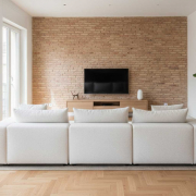 cuanto-cuesta-reforma-madrid-en-2025 La imagen muestra un salón moderno y luminoso en un apartamento de Chamberí, Madrid. Una de las paredes presenta ladrillo visto, contrastando con las paredes blancas restantes. El suelo es de parquet de madera clara en espiga. Un gran sofá modular blanco ocupa el centro de la estancia, orientado hacia un mueble bajo de madera clara con un televisor colgado encima. En la esquina derecha, una planta de interior en una cesta de mimbre aporta un toque de frescura. Grandes ventanas a la izquierda dejan entrar abundante luz natural.