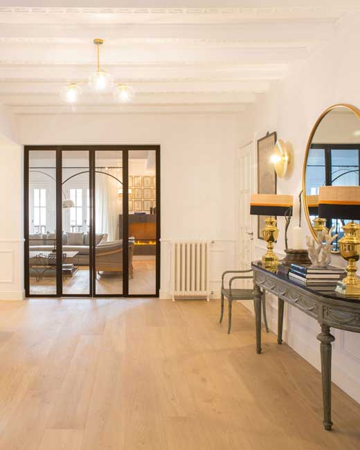 Vista de un recibidor o pasillo elegante de una vivienda reformada en la calle Serrano, un ejemplo de interiorismo en Madrid de estilo clásico renovado. El espacio presenta paredes blancas con molduras y un techo de vigas decorativas blancas. El suelo es de parquet de madera clara. A la izquierda, un cerramiento de cristal y perfilería negra con puertas que reflejan una sala de estar interior. A la derecha, destaca una consola de estilo clásico con patas torneadas, sobre la que reposan dos lámparas doradas con pantallas negras y un gran espejo ovalado enmarcado en oro. También se aprecia un radiador antiguo y una silla auxiliar tapizada de color gris. El ambiente general es de lujo y sofisticación, característico del interiorismo en Madrid en el barrio de Salamanca, especialmente en la calle Serrano.