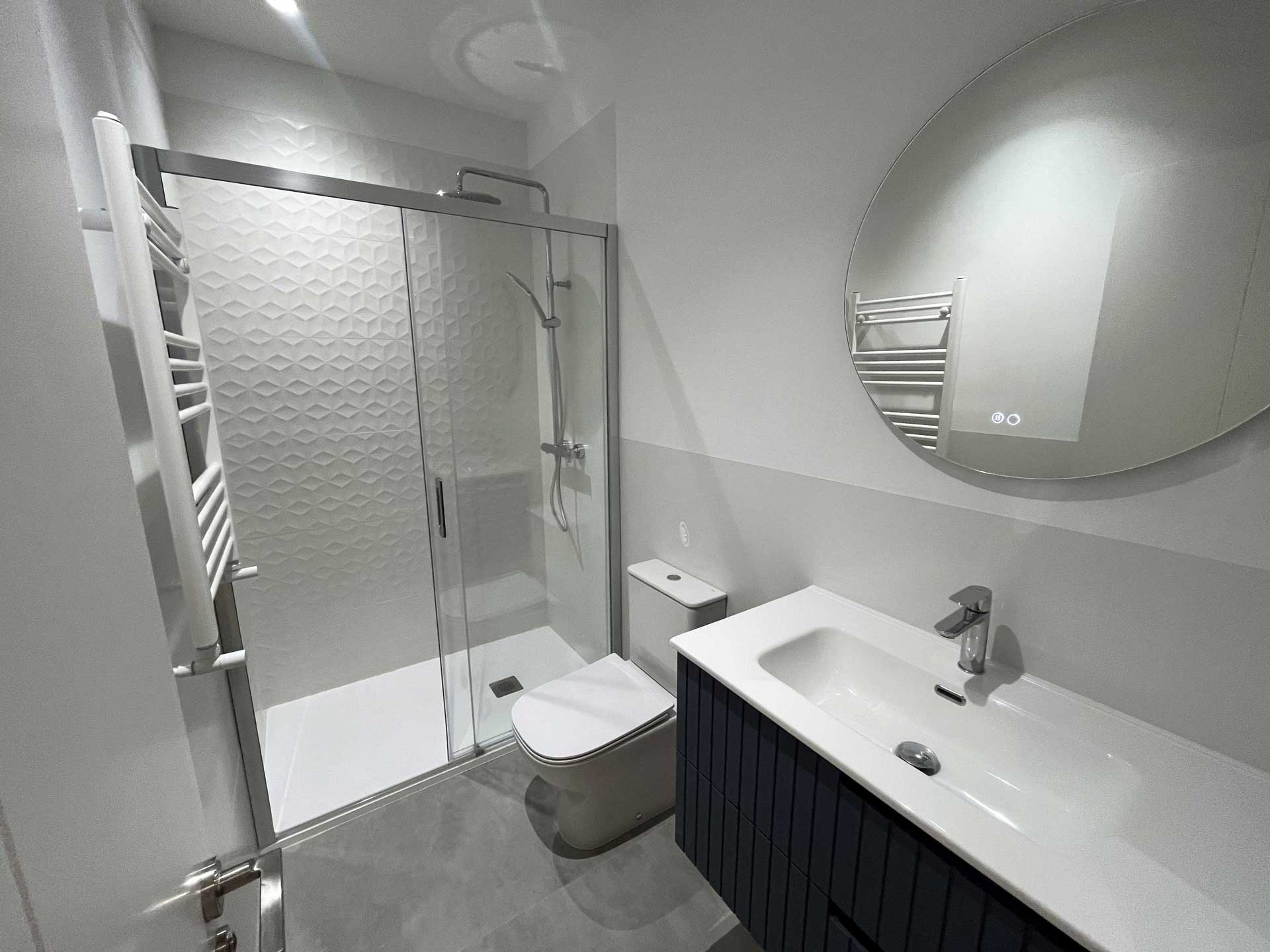 Reforma de baño en Tirso de Molina alicatado hasta media altura, con mueble de madera, espejo redondo y pared de ducha con azulejos decorativos.