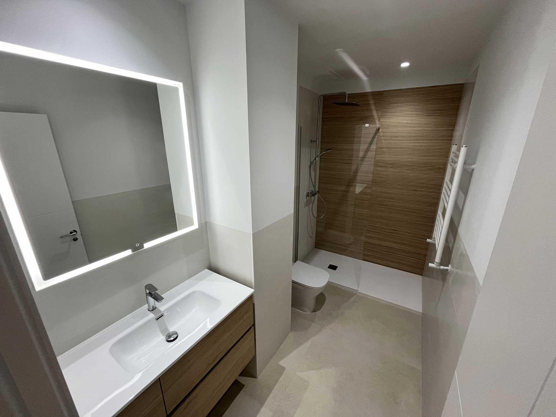 Reforma de baño en vivienda de la calle Tirso de Molina con azulejo hasta media altura, espejo con iluminación LED, mueble de madera, mampara de ducha y pared de ducha en cerámica en habitación madera.