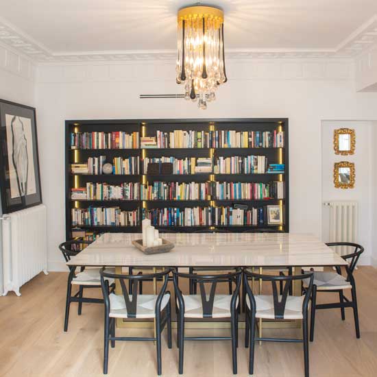 Comedor elegante con gran mesa de mármol, sillas de diseño negras, librería empotrada iluminada y lámpara de techo dorada, reflejando tendencias de decoración para 2026.