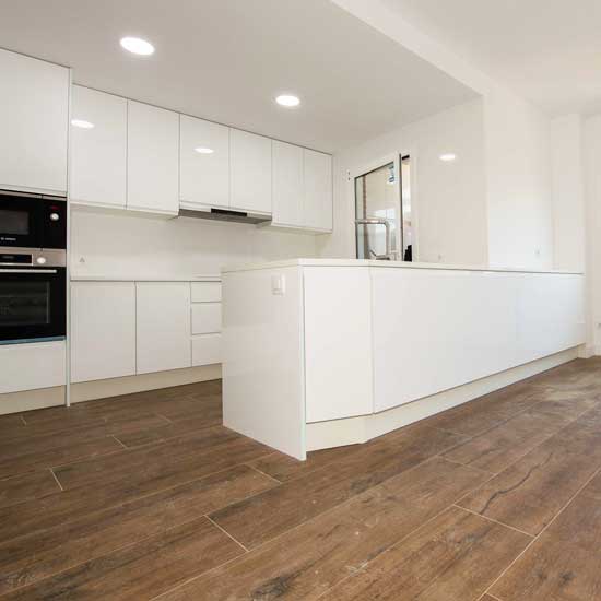 Cocina con península en Madrid de diseño moderno y minimalista, con mobiliario blanco lacado y suelo efecto madera que aporta calidez al espacio