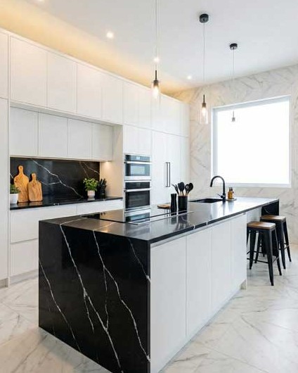 Cocina moderna y lujosa con isla de mármol negro y armarios blancos, diseñada para un proyecto de reformas en Majadahonda. La luz natural de la ventana y la iluminación colgante realzan los acabados de alta gama