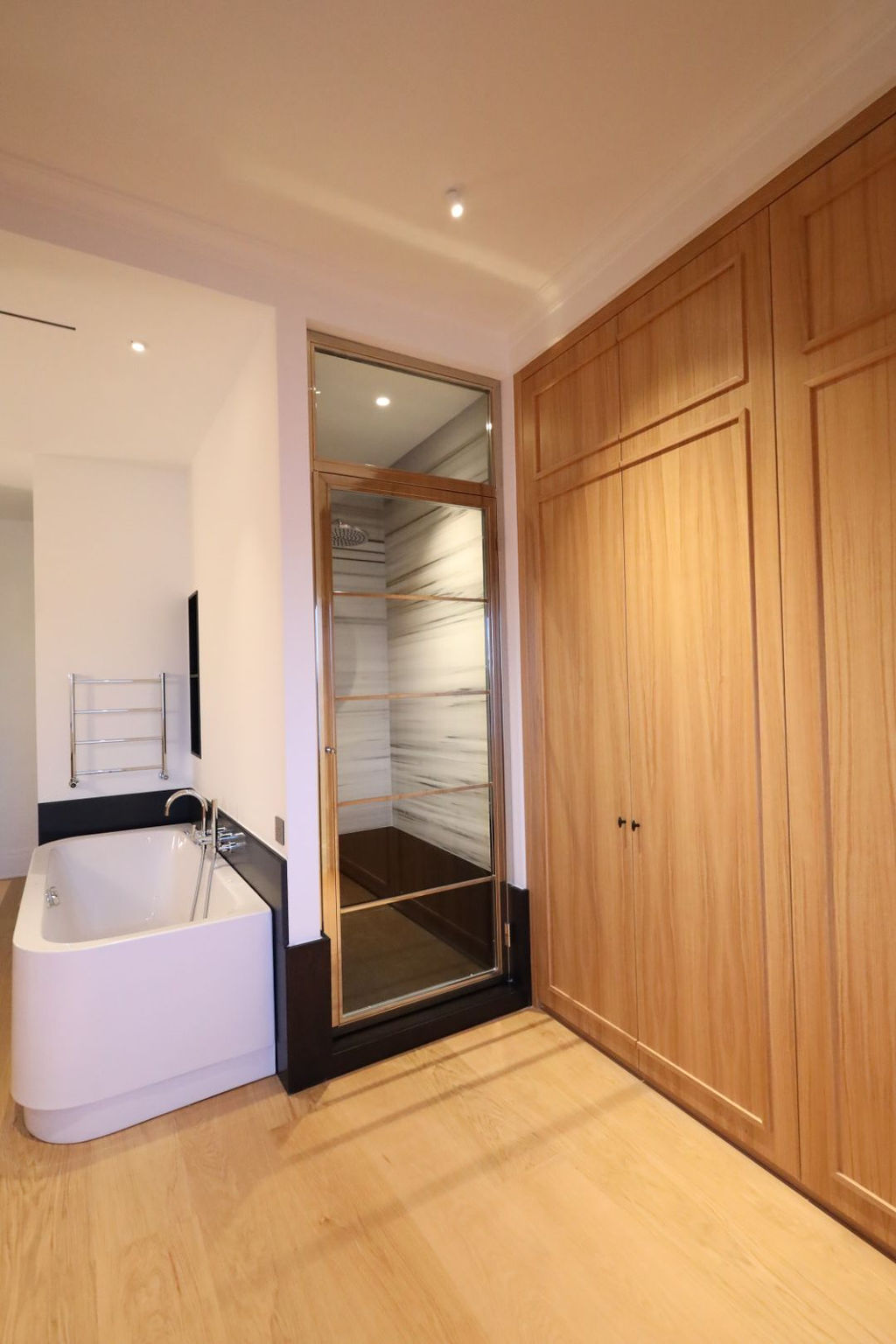 Dormitorio en suite con baño integrado tras una reforma integral en Marqués de Salamanca, con bañera exenta de diseño junto a encimera negra, grifería cromada, mampara de vidrio con perfilería metálica, armarios empotrados de madera a medida y revestimientos en mármol veteado que aportan continuidad visual, elegancia y un ambiente de lujo sereno, todo ello iluminado con luz empotrada que realza los materiales y el diseño contemporáneo del espacio.