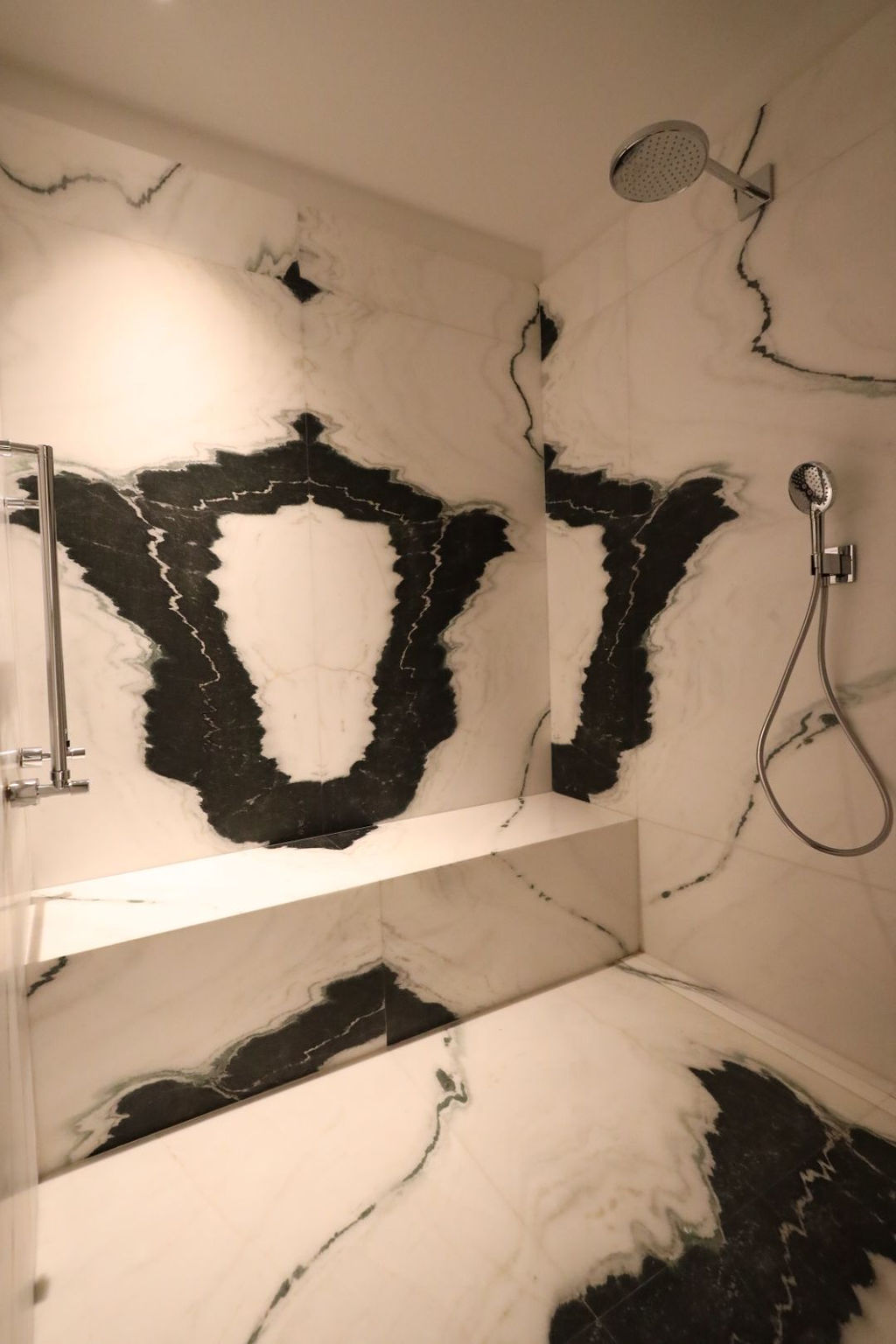Zona de ducha de diseño tras una reforma integral en Marqués de Salamanca, revestida íntegramente en mármol natural de gran formato con vetas contrastadas en blanco y negro, plato de ducha integrado al mismo material, nicho a medida para almacenaje, grifería cromada empotrada con rociador efecto lluvia y mampara de vidrio, creando un espacio elegante, continuo y de alto nivel estético donde el material es el auténtico protagonista.