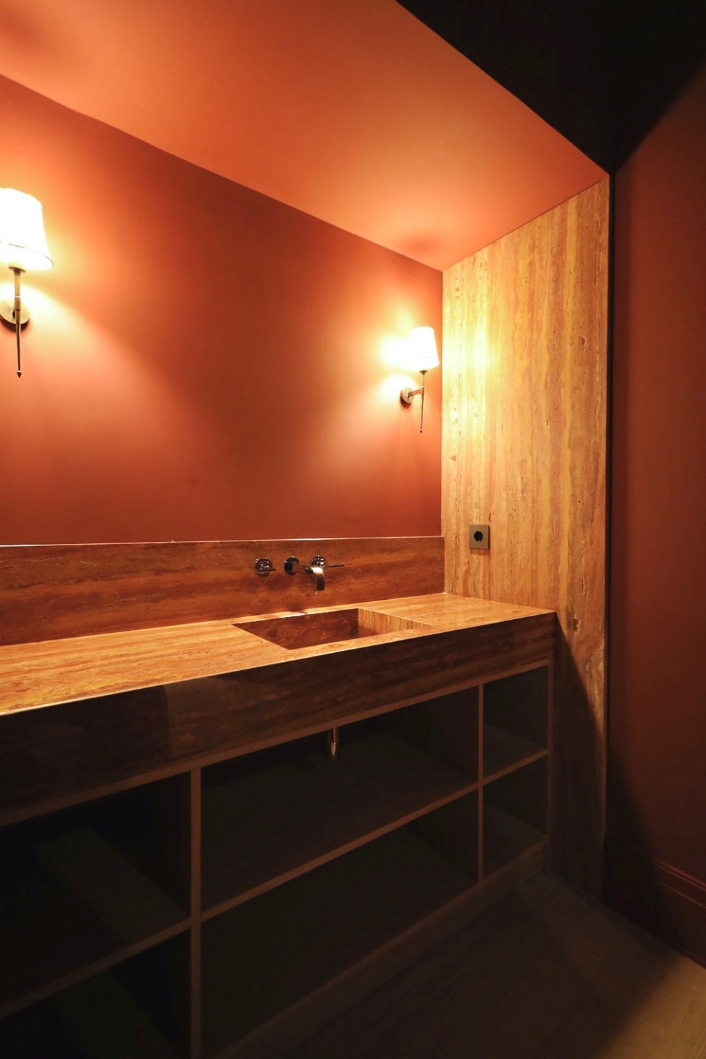 Baño de cortesía de diseño sofisticado tras una reforma integral en Marqués de Salamanca, con lavabo integrado tallado en madera natural, encimera y frontal continuo en acabado pétreo, paredes en tonos cálidos, apliques de iluminación decorativa y grifería empotrada que crean un ambiente íntimo, elegante y de alto impacto estético, donde los materiales nobles y la iluminación ambiental son los protagonistas.