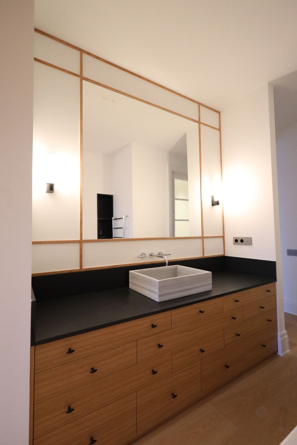 Detalle de baño reformado con diseño elegante y contemporáneo tras una reforma integral en Marqués de Salamanca, con mueble de baño a medida en madera natural con múltiples cajones, encimera negra de acabado mate, lavabo sobre encimera de piedra, grifería mural cromada y gran espejo decorativo con cuarterones de madera que aporta amplitud, luminosidad y sofisticación al espacio, acompañado de iluminación ambiental integrada y paredes en tonos claros que refuerzan la sensación de orden y lujo.
