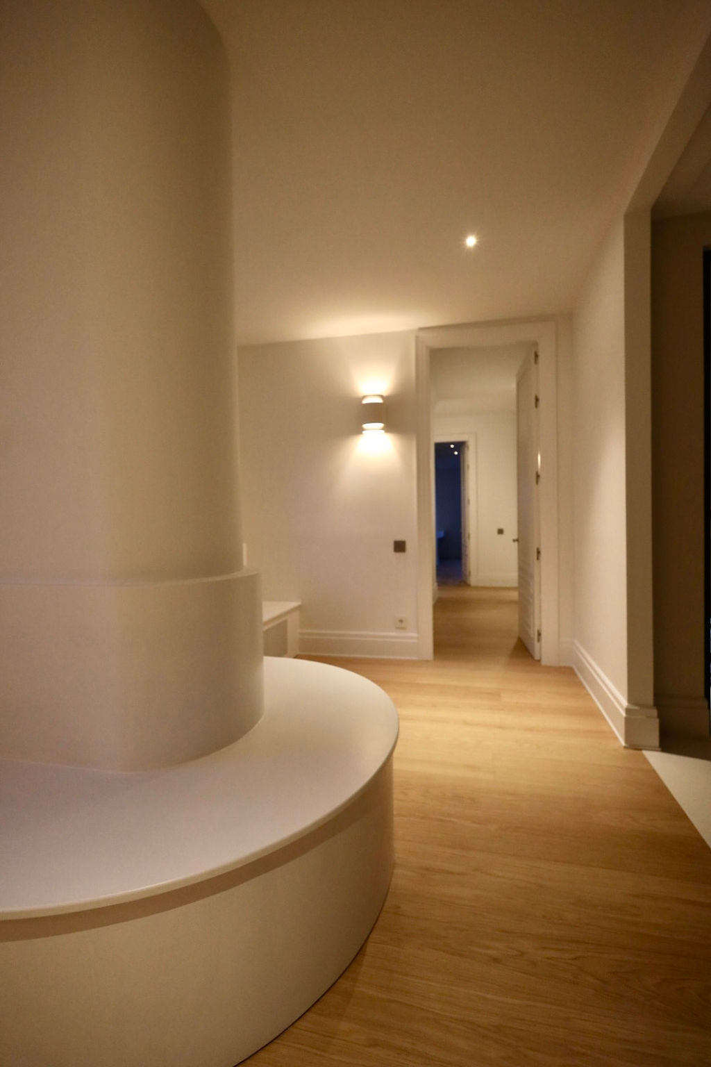 Zona de paso con columna central integrada y banco curvo a medida tras una reforma integral en Marqués de Salamanca, con suelo de madera natural, paredes blancas, iluminación indirecta y diseño contemporáneo que aporta funcionalidad y elegancia al espacio.