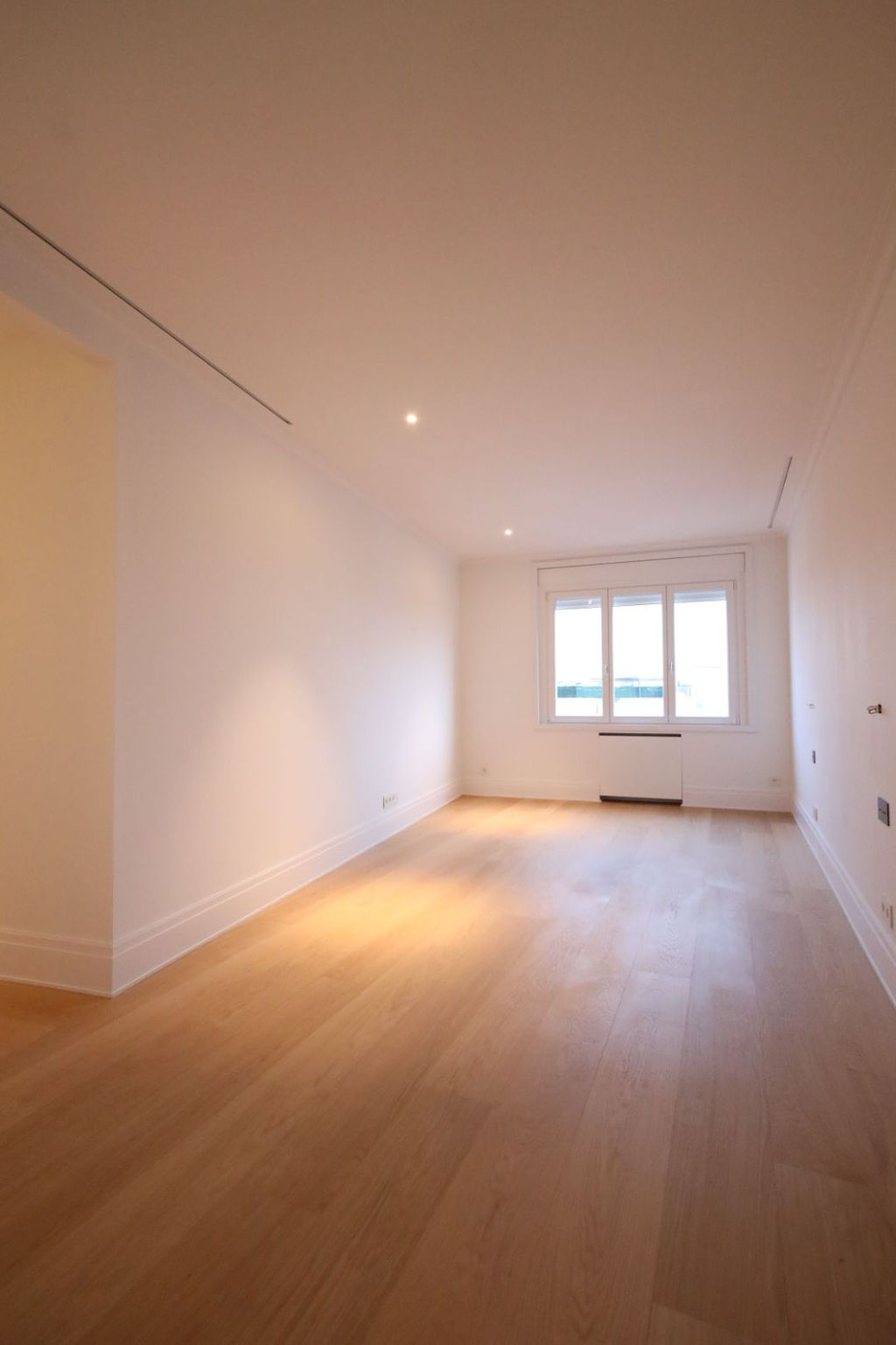 Dormitorio amplio y luminoso tras una reforma integral en Marqués de Salamanca, con suelo de madera natural de gran formato, paredes blancas lisas, iluminación empotrada en techo y gran ventanal que aporta abundante luz natural, creando un espacio sereno, elegante y versátil orientado al confort y al descanso.