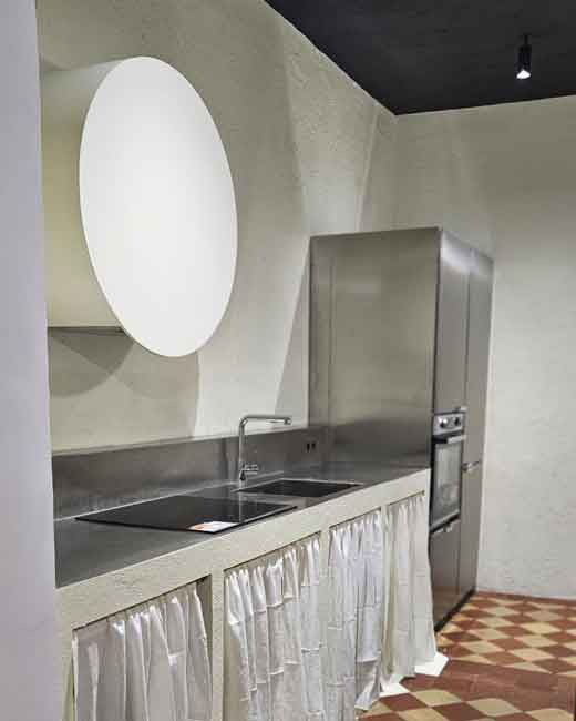 Campana de Filtros y Cocina Industrial en Vivienda de +100 Años en Chueca: Antes y Después. Vista detallada de una cocina moderna con una gran campana de filtros circular retroiluminada en una vivienda de más de 100 años en Chueca, Madrid. Encimeras de acero inoxidable, placa de cocción negra integrada y cortinas de lino blanco cubriendo los armarios inferiores sobre un suelo de baldosas hidráulicas originales restauradas. Esta imagen documenta el resultado del proyecto de reforma 'antes y después'.