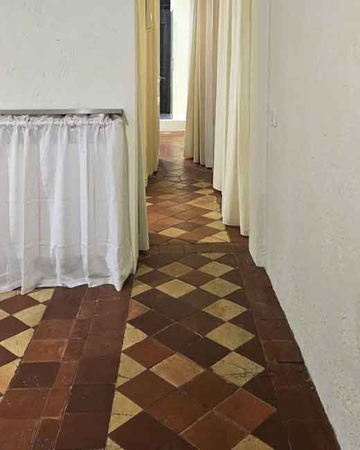 Suelo hidráulico restaurado en vivienda de más de 100 años en Chueca: Antes y Después. Un pasillo interior reformado en una vivienda de más de 100 años en Chueca, Madrid, destacando el suelo hidráulico de mosaico centenario restaurado con patrón de diamantes de colores marrón y ocre. Las paredes son de estuco blanco texturizado con cortinas ligeras en las puertas. Esta imagen documenta el resultado del proyecto de reforma 'antes y después'.