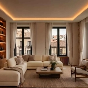 Acogedor salón moderno diseñado con principios de neuroarquitectura en reformas en Madrid, con un gran sofá seccional beige, estantería de madera integrada e iluminada, paredes de piedra texturizada y grandes ventanas con vistas a la ciudad.