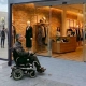 Persona en silla de ruedas eléctrica accediendo por una rampa interior perfectamente enrasada a una boutique moderna en planta baja con puertas automáticas acristaladas, proyecto de reforma de local comercial ejecutable mediante las ayudas del Plan Adapta 2026.