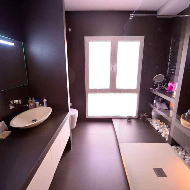 Baño suite negro blanco - Reforma en calle Goya 15 años después Vista angular del cuarto de baño de la suite principal perteneciente al proyecto de reforma en la calle Goya, fotografiado 15 años después. Se aprecia la encimera oscura con lavabo tipo bol, el suelo continuo de resina o microcemento, el inodoro suspendido al fondo y una zona de ducha espaciosa con ventana al patio, iluminada por luz natural filtrada.