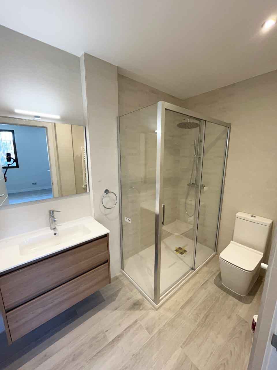 oficina-ciudad-barcelona-madrid-convertida-dos-viviendas-bano-piso-uno Vista general del baño del Piso Uno. Incluye plato de ducha con mampara de vidrio, grifería empotrada, mueble suspendido con cajones texturizados en madera y espejo iluminado. Ejemplo de acabados de alta calidad en la transformación de oficina en Pacífico convertida en dos viviendas.