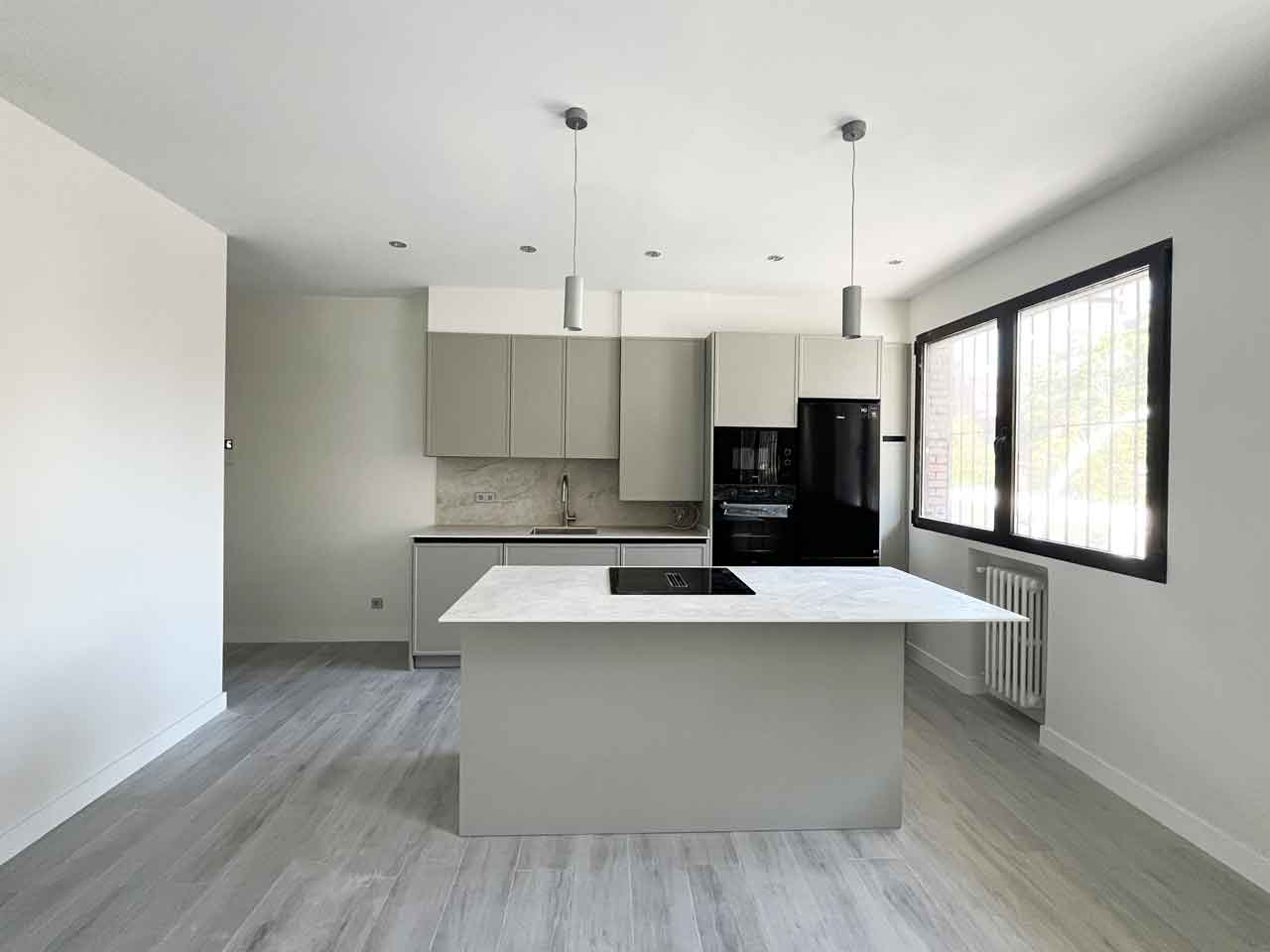 oficina-ciudad-barcelona-madrid-convertida-dos-viviendas-concepto-abierto-piso-uno Amplia cocina del Piso Uno. Destaca la isla central con placa de cocción, muebles de columna con electrodomésticos integrados en negro y gris, y encimera efecto mármol. Espacio luminoso gracias a los ventanales con rejas de seguridad, perfecto para la oficina en Pacífico convertida en dos viviendas.