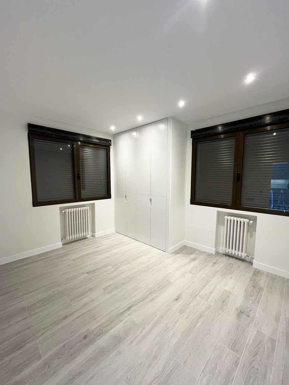 oficina-ciudad-barcelona-madrid-convertida-dos-viviendas-dormitorio-principal-piso-uno Vista del dormitorio principal delo piso uno de la oficina en Pacífico convertida en dos viviendas. Cuenta con dos ventanas oscuras con persianas, dos radiadores de hierro fundido restaurados y un armario empotrado blanco de cuatro puertas. Suelo de tarima en espiga gris.