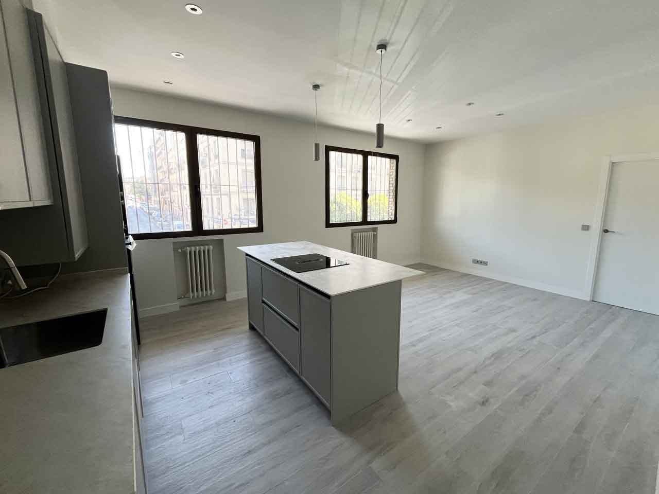 oficina-ciudad-barcelona-madrid-convertida-dos-viviendas-salon-cocina-americana-piso-uno Panorámica del área social del Piso Uno. Muestra la integración de la cocina gris con isla y encimera blanca en un salón diáfano y luminoso. Se observan los ventanales con rejas, radiadores y focos empotrados en el techo. Resultado final de una oficina en Pacífico convertida en dos viviendas.