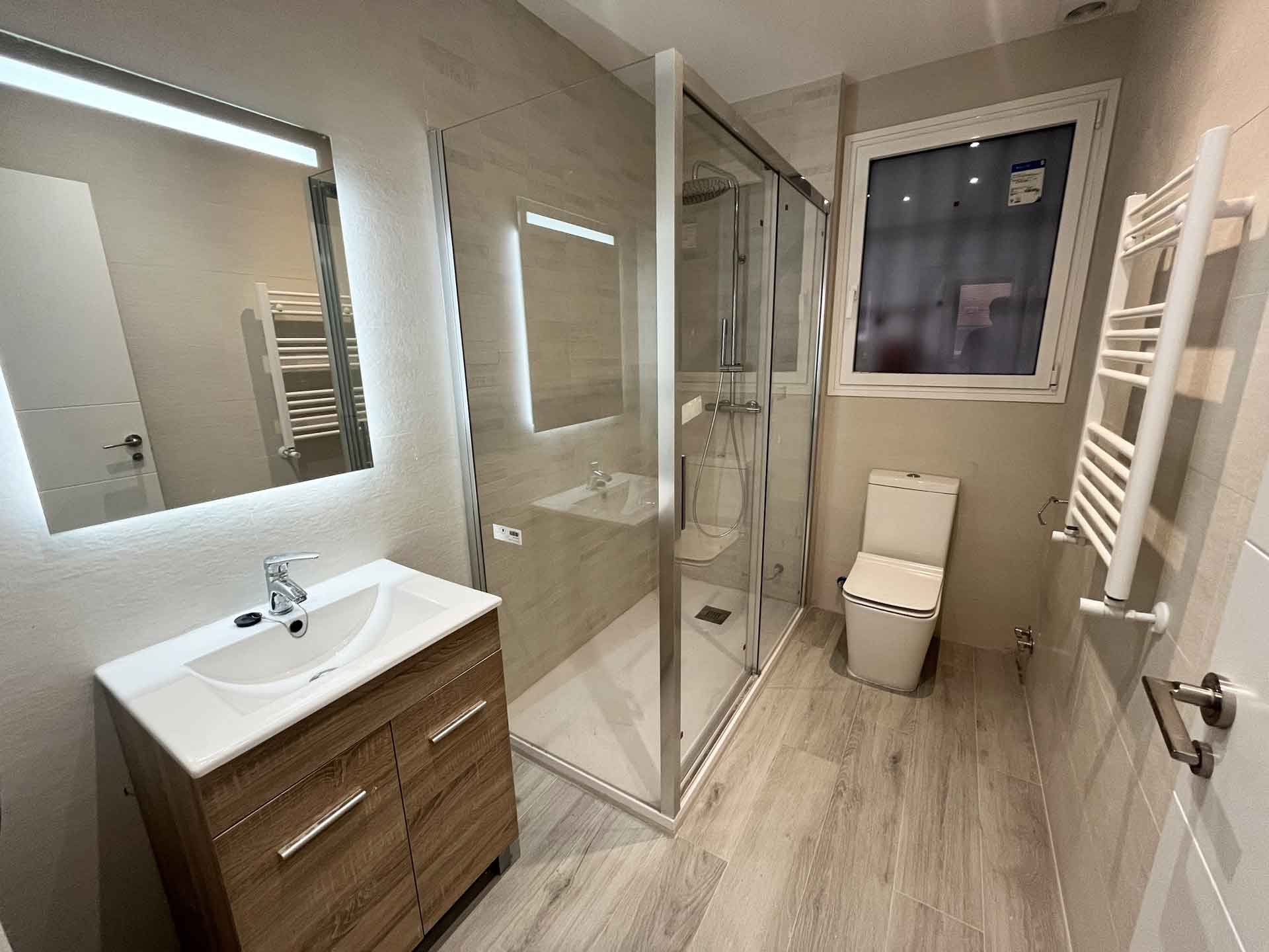 oficina-madrid-convertida-dos-viviendas-bano-piso-dos Baño completo del Piso Dos dentro de la reforma de una oficina convertida en dos viviendas en Madrid. Equipado con mueble de baño en acabado madera con lavabo cerámico, espejo retroiluminado LED, mampara de ducha de cristal con perfiles de aluminio, toallero radiador blanco y ventana con luz natural.