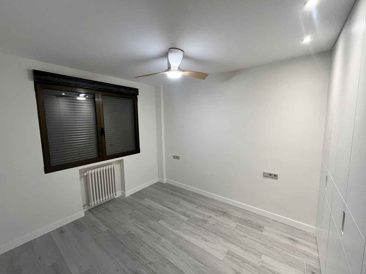 oficina-madrid-convertida-dos-viviendas-salon-piso-dos Vista amplia del dormitorio principal del Piso Dos. Cuenta con carpintería exterior oscilobatiente en tono oscuro con persiana, radiador de hierro restaurado, ventilador de techo y armario empotrado a juego con la tarima gris. Forma parte de la obra de oficina en Pacífico convertida en dos viviendas