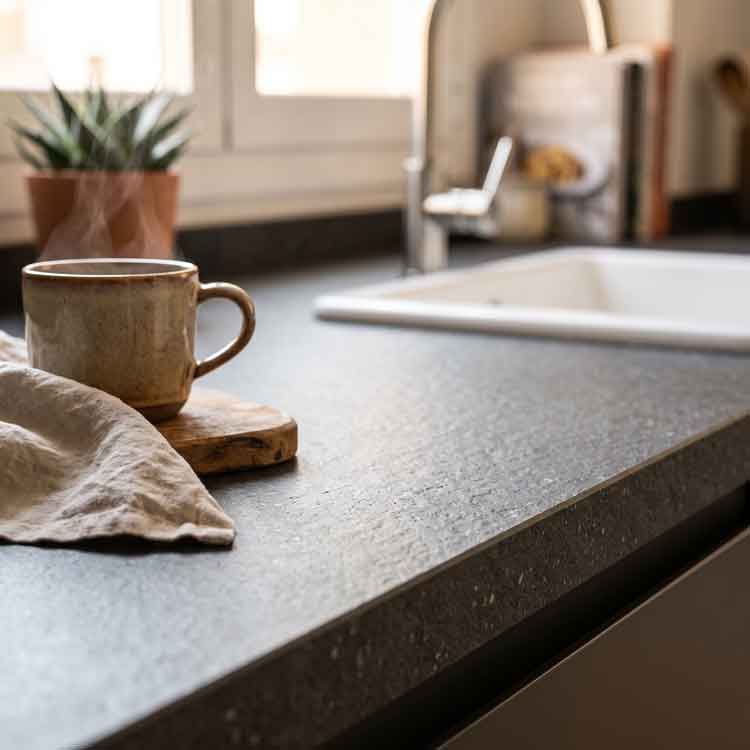 Primer plano de una encimera de piedra sinterizada en color oscuro, luz natural y una taza de cerámica. Este recurso visual complementa nuestra comparativa entre encimeras de cuarzo versus encimeras de piedra sintetizada, tendencia en 2026 para proyectos de interiorismo y reformas integrales de cocina.